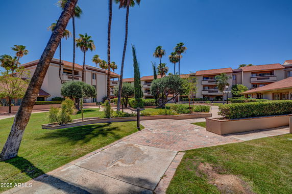 Front view of a residential complex with multiple three-story buildings featuring balconies and surrounded by landscaped gardens and tall palm trees.