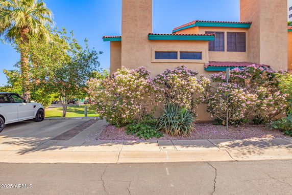 Front view of a house with light brown exterior walls and a tiled roof surrounded by flowering bushes.