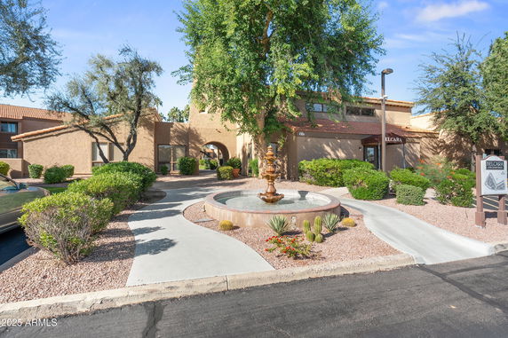 Front view of a two-story building with an entrance path, surrounded by trees and shrubs, featuring a fountain in the foreground.