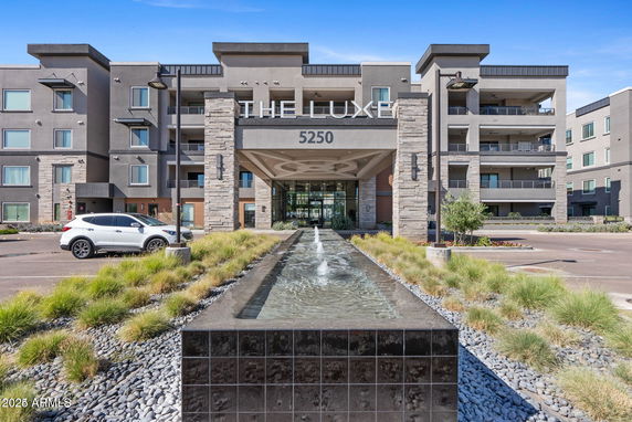 Front view of a modern multi-story building with balconies and a central entrance, featuring a water fountain in the foreground.