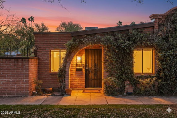 Front view of a single-story brick house with an arched entrance and illuminated windows during sunset.