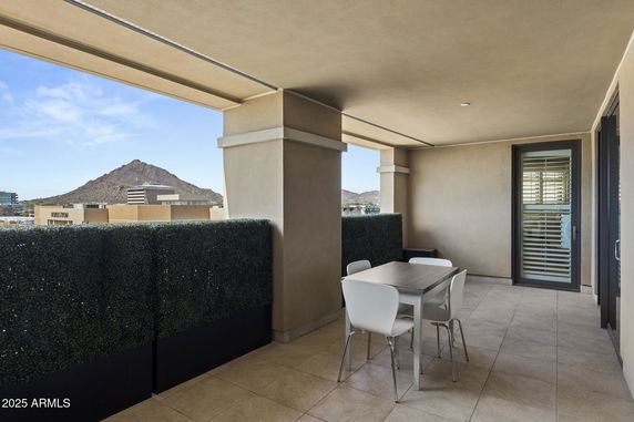 Covered balcony area with table and chairs offering a view of a mountain and surrounding buildings.