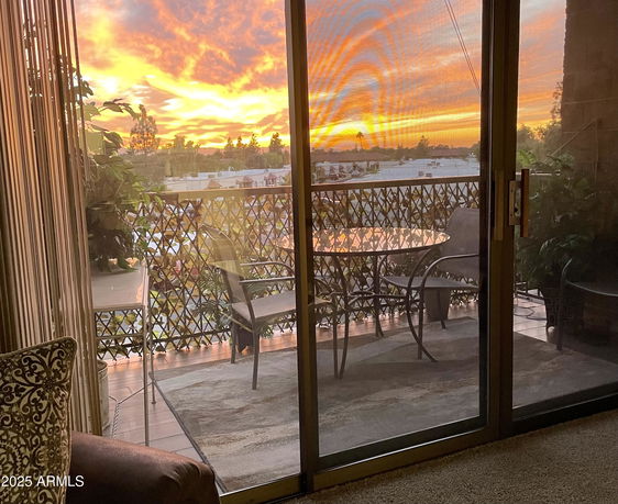 View of a balcony with outdoor furniture and sunset in the background through a glass door.