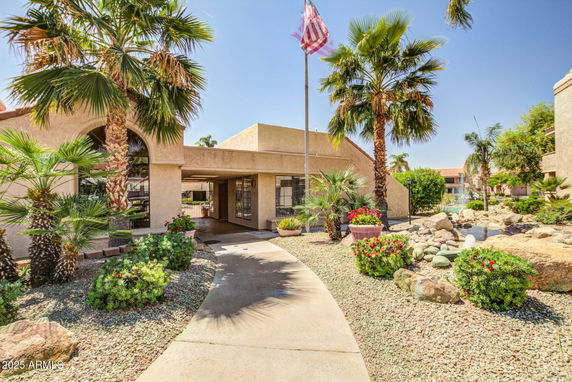 Front view of a building with a covered entrance and a flagpole, surrounded by palm trees and landscaped garden.