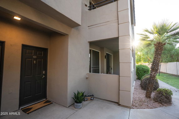 Front view of a building entrance with a balcony above and a door with a mat and potted plant.