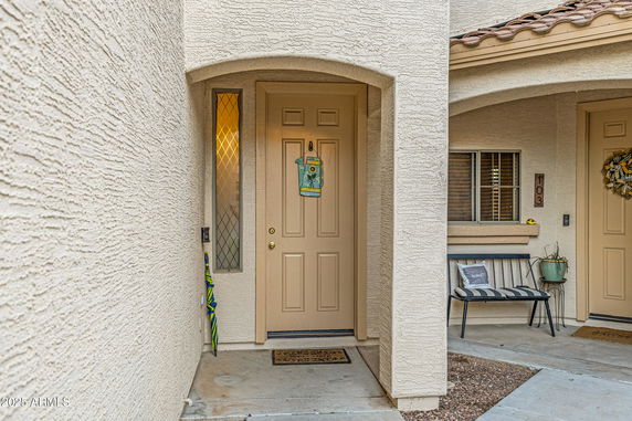 Front view of a house entrance with a beige door and a small window beside it, featuring a welcome mat and a nearby bench.