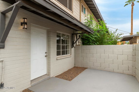 Entrance to a building with a white door and window, surrounded by a small concrete patio and brick wall.