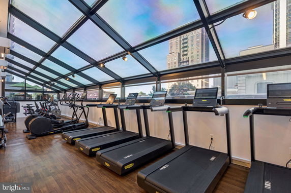 Indoor gym area with treadmills and exercise equipment, featuring a glass ceiling and views of tall buildings.