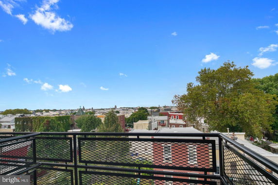 Elevated view of cityscape from a balcony with metal railing, showing rooftops and trees.