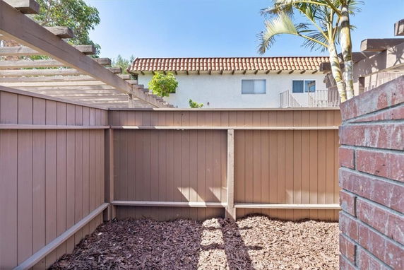 View of a fenced outdoor area with a wooden pergola and a house in the background.