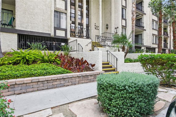 Front view of a multi-story residential building with stairs leading to the entrance and surrounding plants.