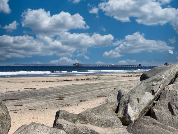 View of a beach with a pier extending into the ocean under a partly cloudy sky.