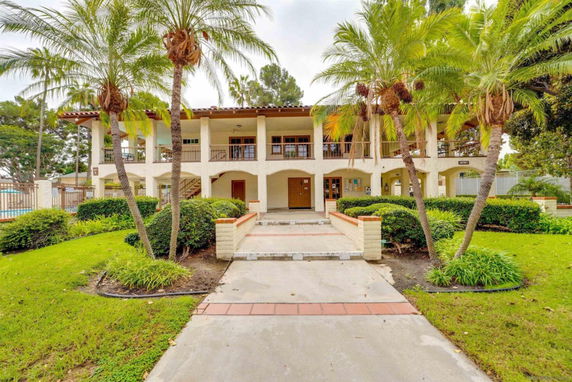 Front view of a two-story building with arches and balconies, surrounded by palm trees and shrubs.