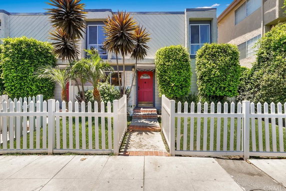Front view of a house with a red door, white picket fence, and decorative plants.