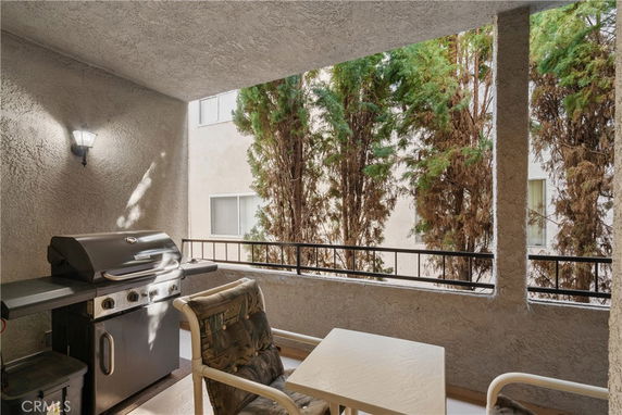 Covered outdoor patio with a grill and seating area, partially enclosed by walls with views of neighboring building and trees.