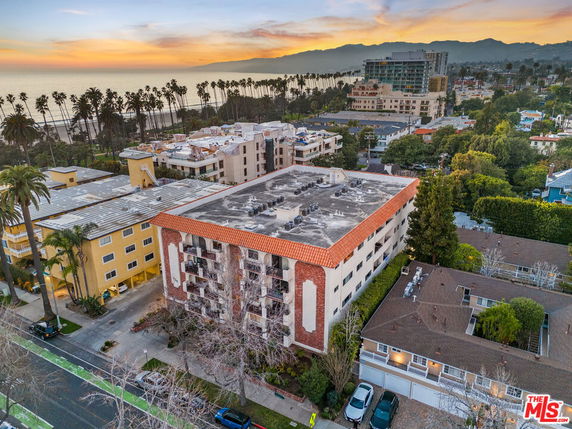 Aerial view of a residential area with an apartment building in the foreground and a coastline with palm trees and ocean in the background.
