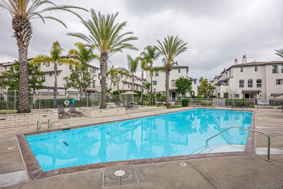 Outdoor swimming pool in a residential complex with palm trees and multi-story buildings around.