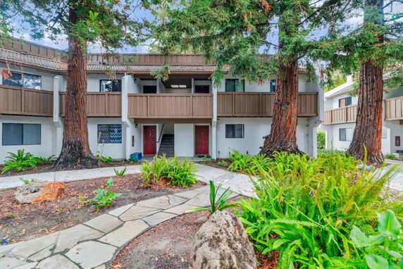 Front view of a two-story apartment building with wooden balconies and surrounded by large trees.