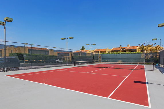 Tennis court with red surface and surrounding fence, located near residential buildings.