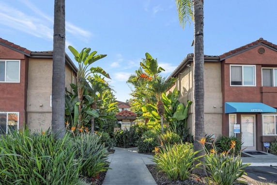 View of a two-story building with brown and beige exterior, featuring a pathway between palm trees and lush greenery.
