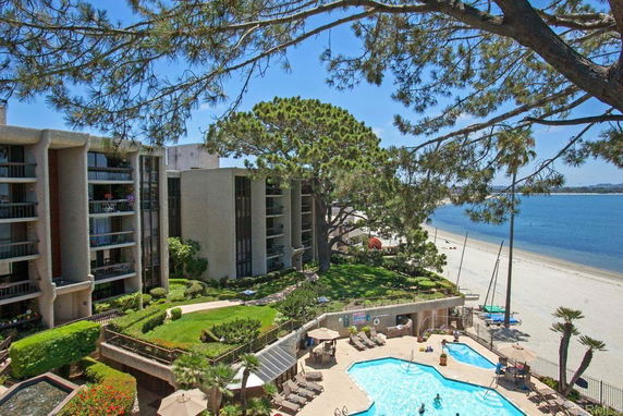 View of a multi-story building near a beach with a swimming pool and surrounding trees.