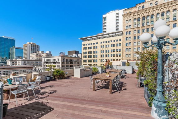 Rooftop area with seating, tables, and decorative plants, surrounded by urban buildings.
