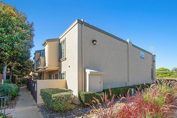 Side view of a two-story residential building with beige exterior walls and green plants surrounding the pathway.