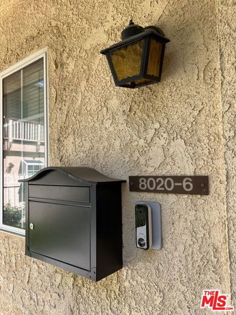 Exterior wall of a house with a window, wall-mounted black mailbox, doorbell camera, and an outdoor light fixture.