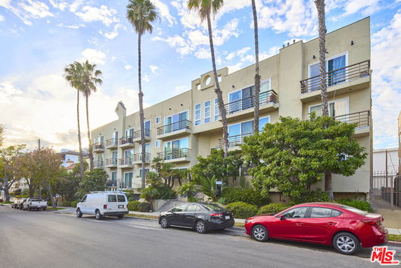 Front view of a multi-story apartment building with balconies and palm trees lining the sidewalk.