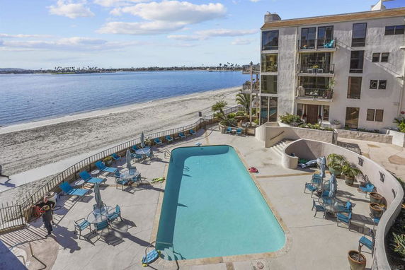 View of an outdoor swimming pool area adjacent to a sandy beach and a multi-story building.