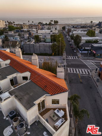 Aerial view of a rooftop with patio furniture and a red-tiled roof, overlooking a street and the ocean.