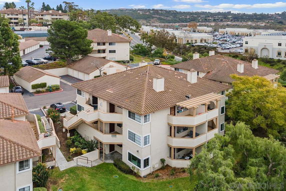 Elevated view of a residential complex with brown tile roofs and surrounding vegetation, facing a parking area and hills in the background.