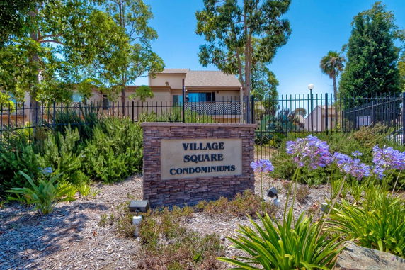 Front view of a condominium building with a sign reading 'Village Square Condominiums' surrounded by landscaping.