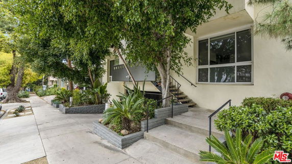 Front view of a building with stairs and large windows, surrounded by trees and plants.