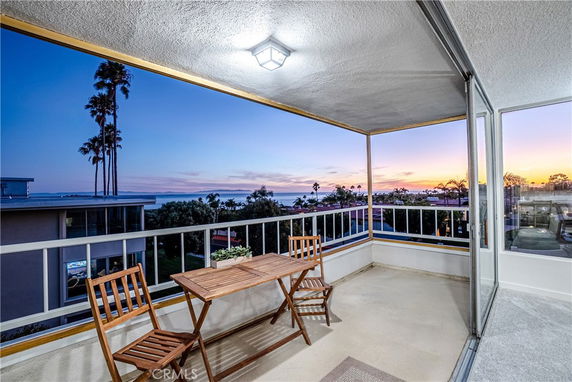 Balcony with wooden table and chairs overlooking a coastal view with palm trees and sunset.