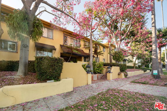 Front view of a two-story residential building with yellow walls and awnings, surrounded by pink flowering trees.