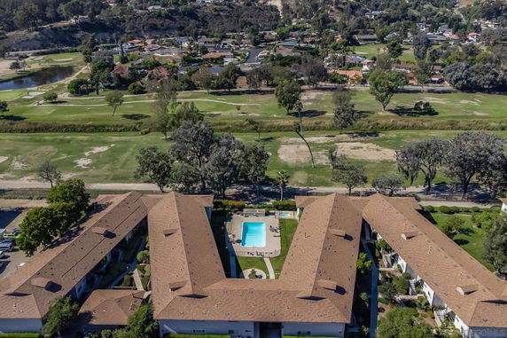 Arial view showcasing a residential complex with a swimming pool in the center, surrounded by greenery and distant houses.