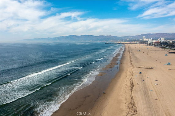Panoramic view of a long sandy beach and ocean waves with mountains and buildings in the distance.