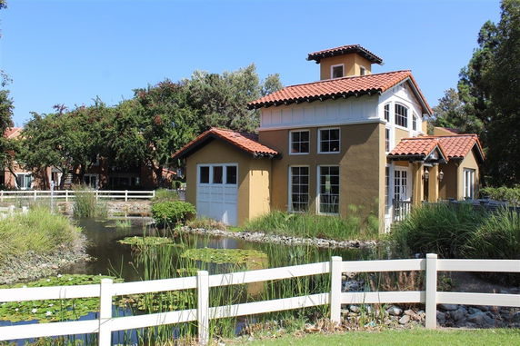 Front view of a house with a red tile roof and multiple windows, surrounded by a small pond and greenery.