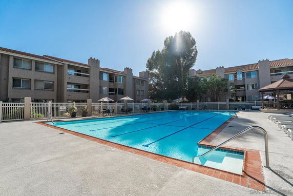 Outdoor swimming pool area with sun loungers and a surrounding fence, adjacent to multi-story residential buildings.