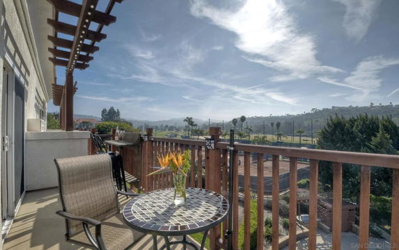 Balcony with chairs and a table overlooking a tennis court and scenic hills.