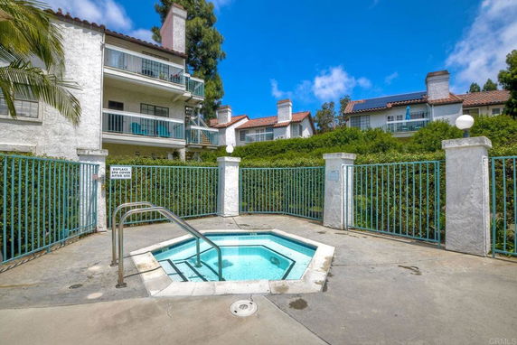 Outdoor view featuring a small hot tub surrounded by a gated fence, with multi-story residential buildings in the background.
