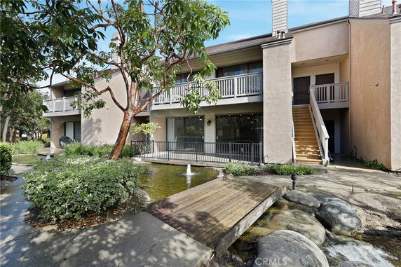 Front view of a two-story building with balconies and a wooden footbridge over a small pond.