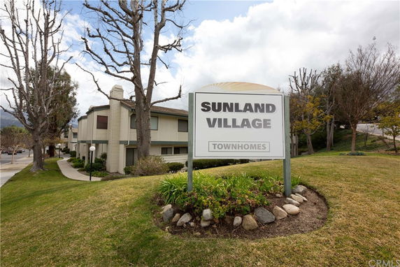 Front view of a two-story townhome building with a white exterior and a sign labeled 'SUNLAND VILLAGE TOWNHOMES'.