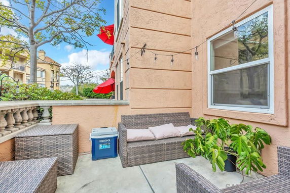 Outdoor seating area with wicker furniture and potted plant adjacent to a building, red umbrellas visible in background.