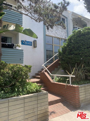 Front view of a multi-story building with stairs leading to the entrance and plants along the pathway.