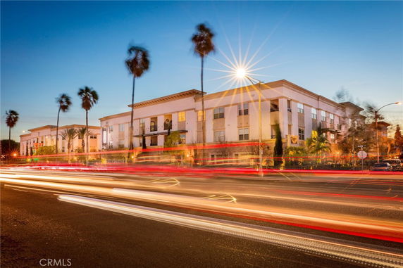 Front view of a multi-story apartment building with tall palm trees and light trails from passing cars.