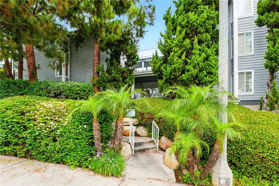 Front view of a building with grey siding and lush greenery, including palm trees and shrubs.