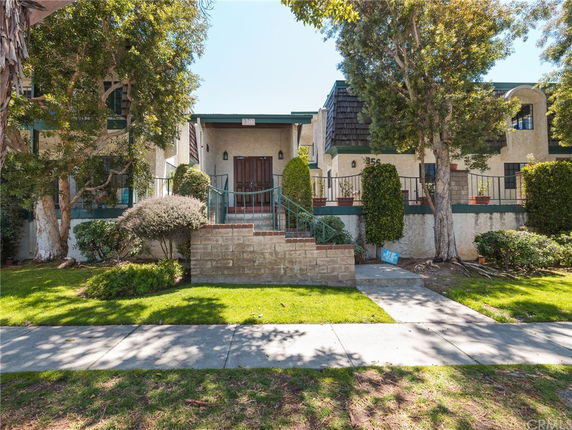 Front view of a house with a raised entrance and stairs, surrounded by trees and shrubs.