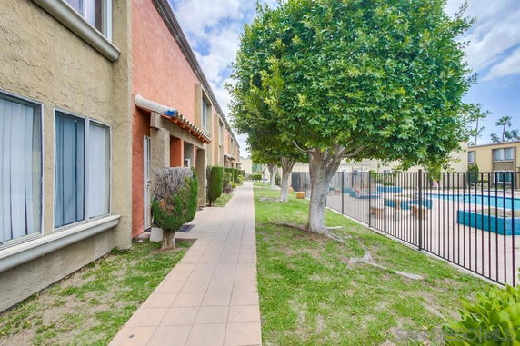View of a pathway next to a residential building with trees and a fenced swimming pool area.
